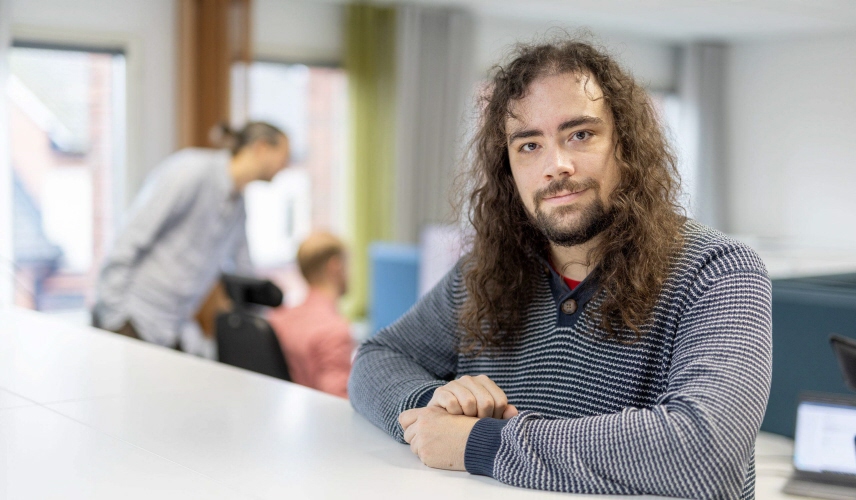 Man lutar sig mot ett bord och tittar in i kameran, kollegor i bakgrunden.