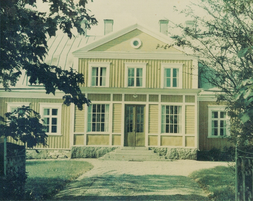 Bleknat fotografi av pampigt gult trähus med vita knutar och grön glasinfattad entrédörr, med bred grusgång som leder fram till huset samt del av stenstolpe och prunkande träd i förgrunden